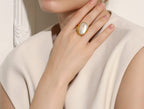 Hand wearing a gold ring with a large gemstone, against a neutral background